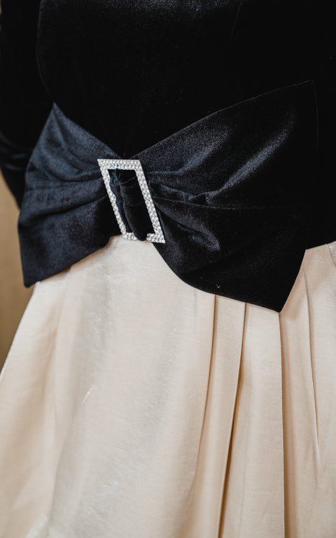 Long sleeve, black dress has ribbon belt and beige, satin bow swing skirt.